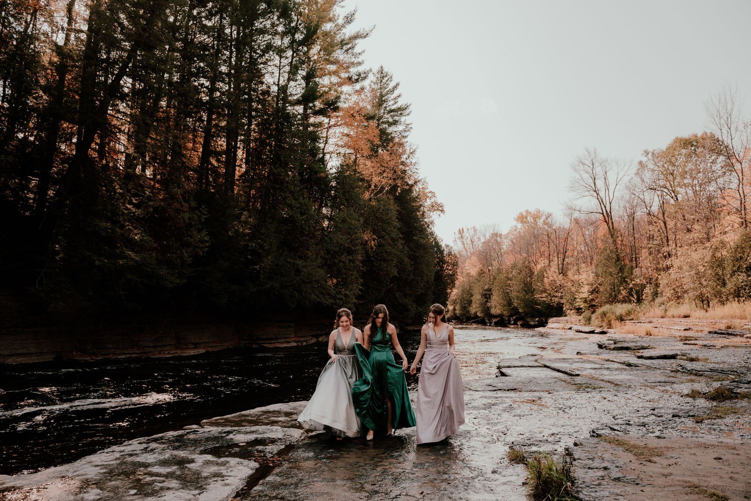 Three people in formal wear stand together by a river in autumn.