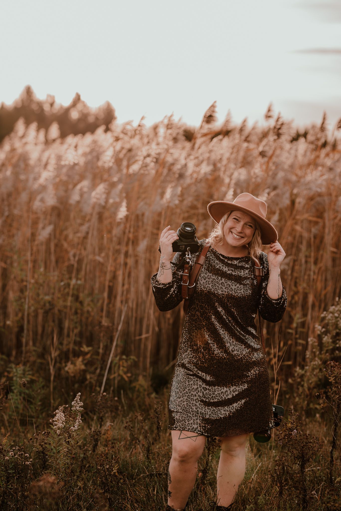 Woman in leopard print dress and hat poses in front of tall grass at sunset.