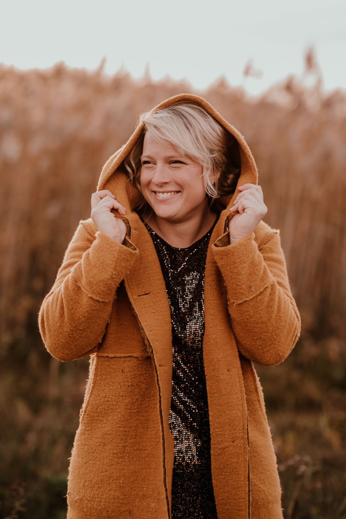 Woman wearing orange coat and sequin top smiles while pulling up hood in autumn field.