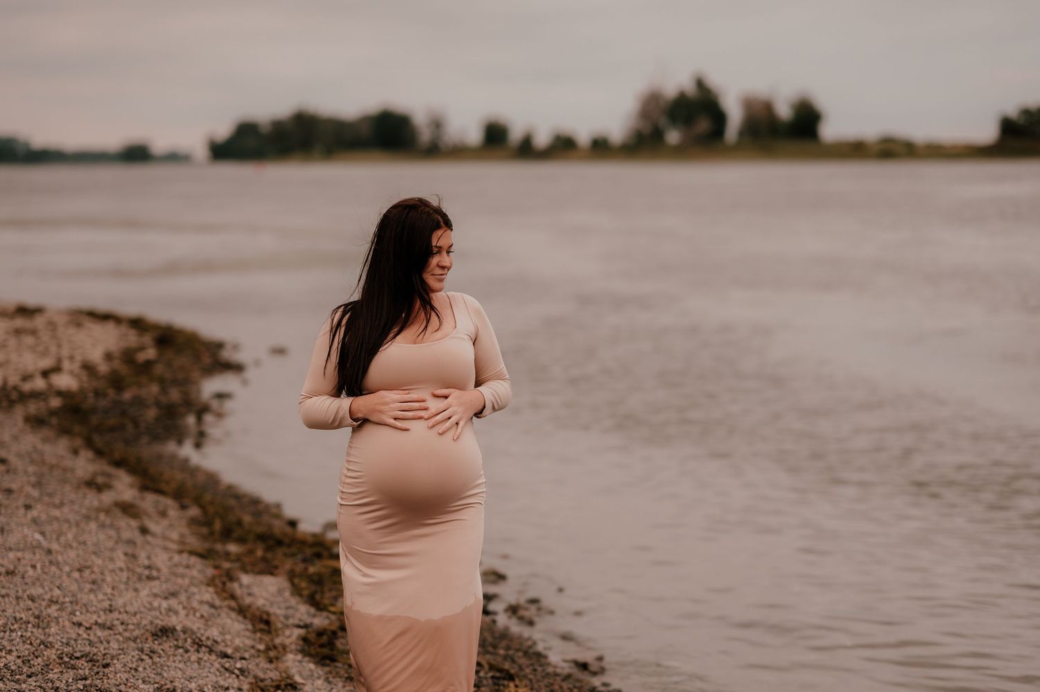 Woman in pink dress stands on shoreline at sunset.