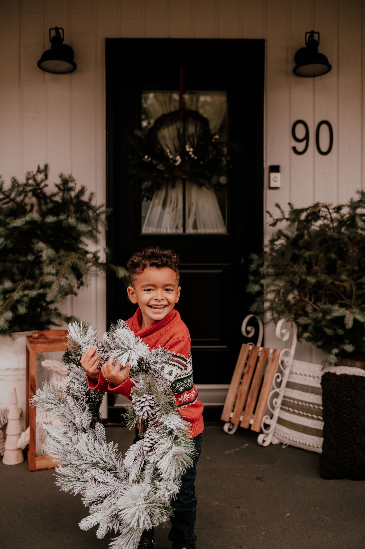 Joyful child in red holding a frosted Christmas wreath on a porch with black door and house number 90.