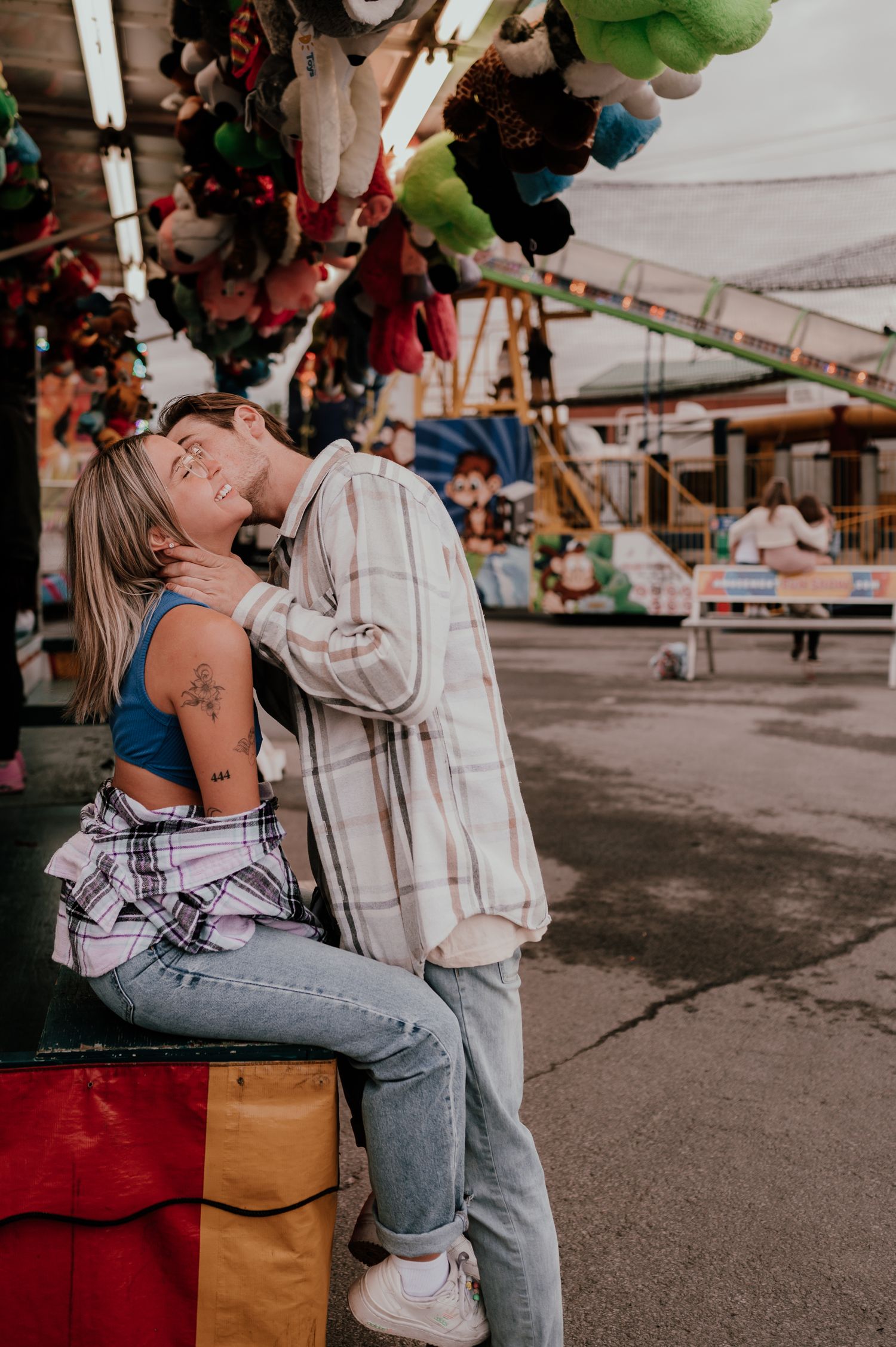 Couple sharing a romantic moment at carnival with colorful decorations and rides in background.