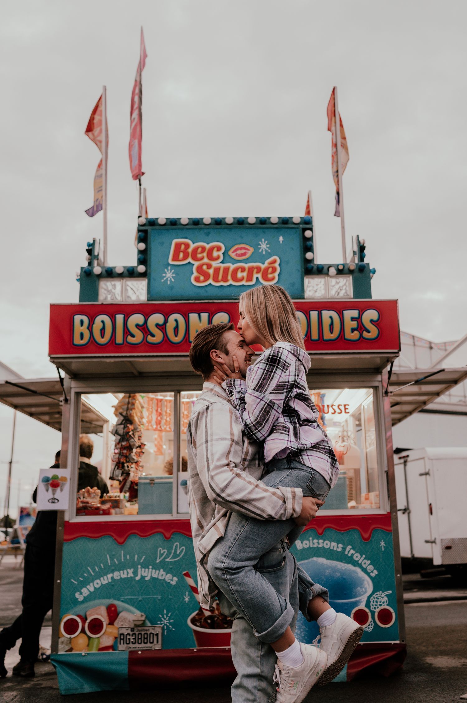 Couple embracing in front of vintage carnival snack stand with French text.