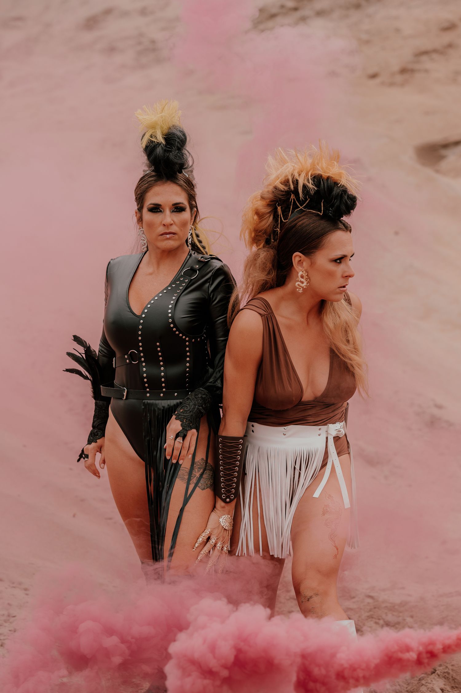 Two fashionable figures pose in edgy swimwear outfits surrounded by pink smoke in a desert setting.