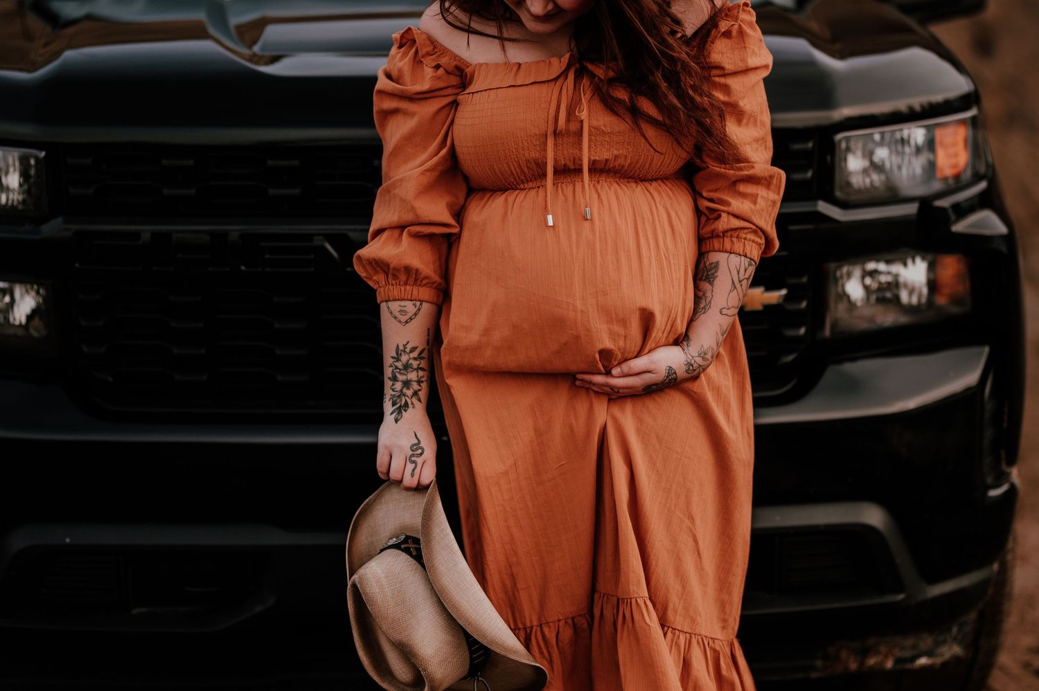 Person in flowing orange maternity dress poses beside black vehicle.