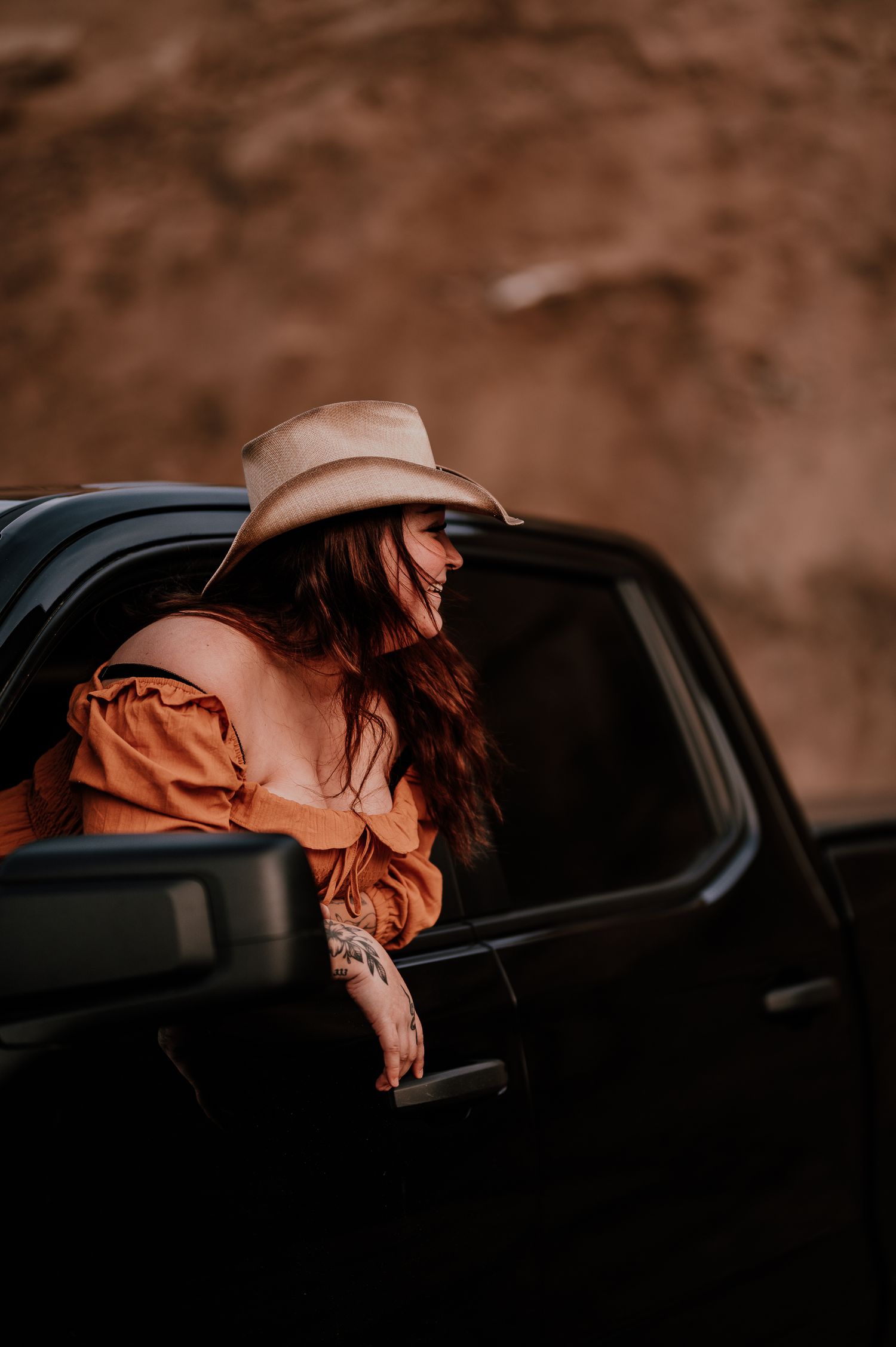 Person wearing cowboy hat leans out of black vehicle window in warm sunset lighting.