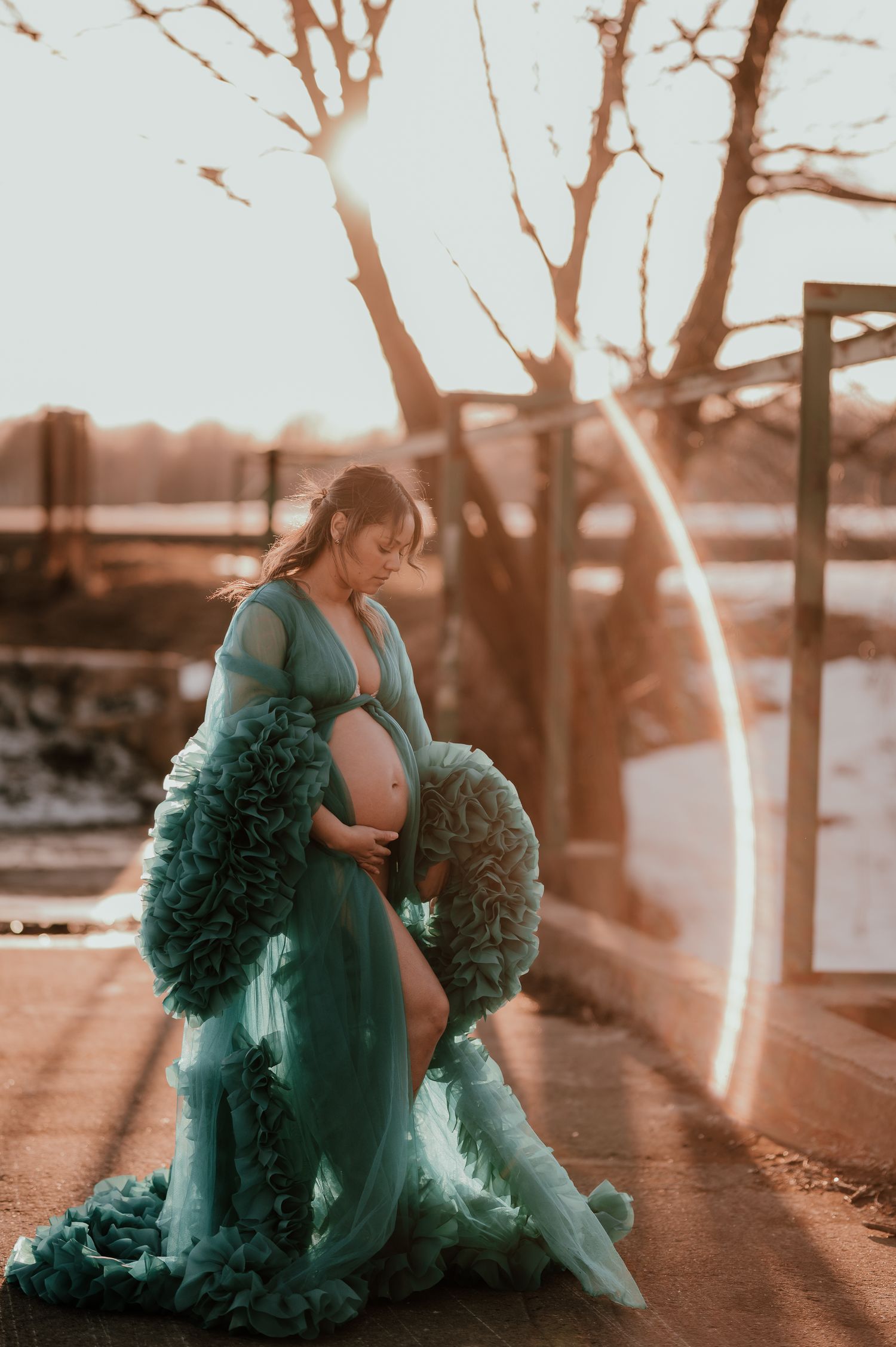 Figure in elegant teal maternity gown poses during sunset with dramatic fabric details.