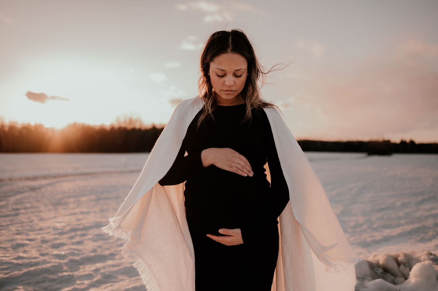 A pregnant woman in black dress and white cardigan stands in a snowy field at sunset.