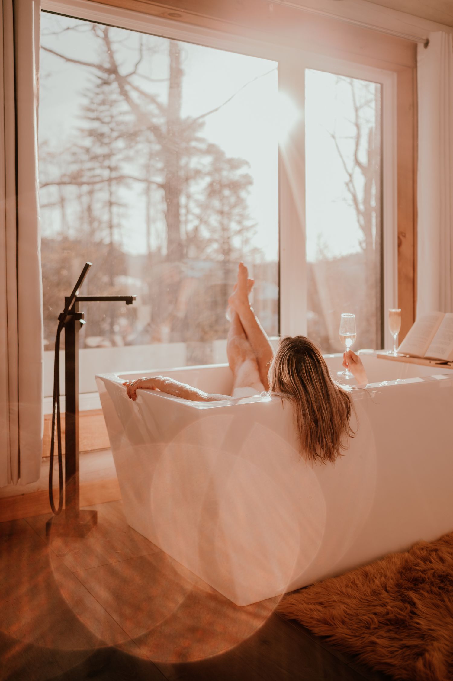 Woman relaxing in modern freestanding bathtub with forest view through large windows.