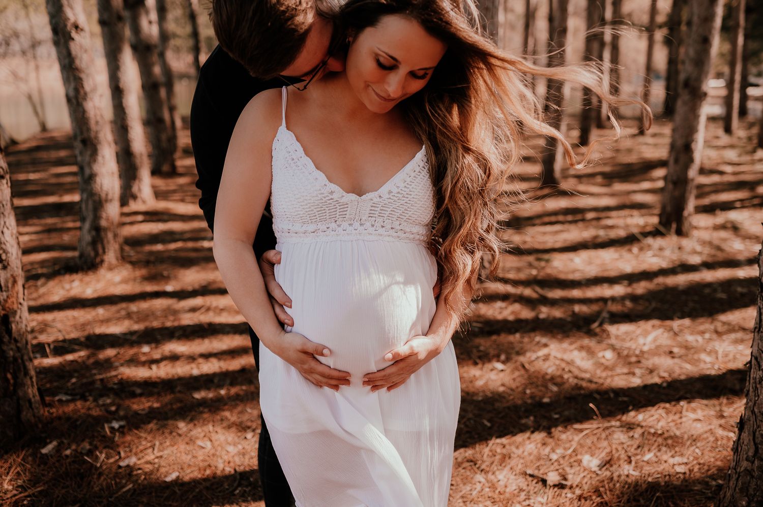 Maternity photo of woman in white lace dress touching belly in sunlit forest.