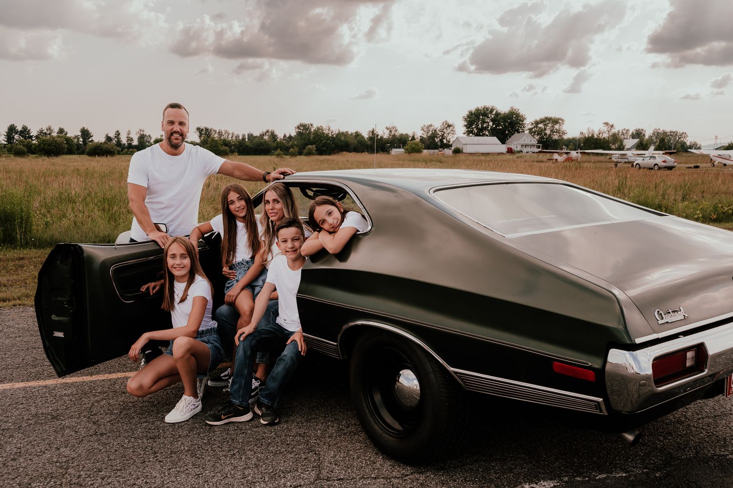 Classic black muscle car on rural road with family posed beside it.