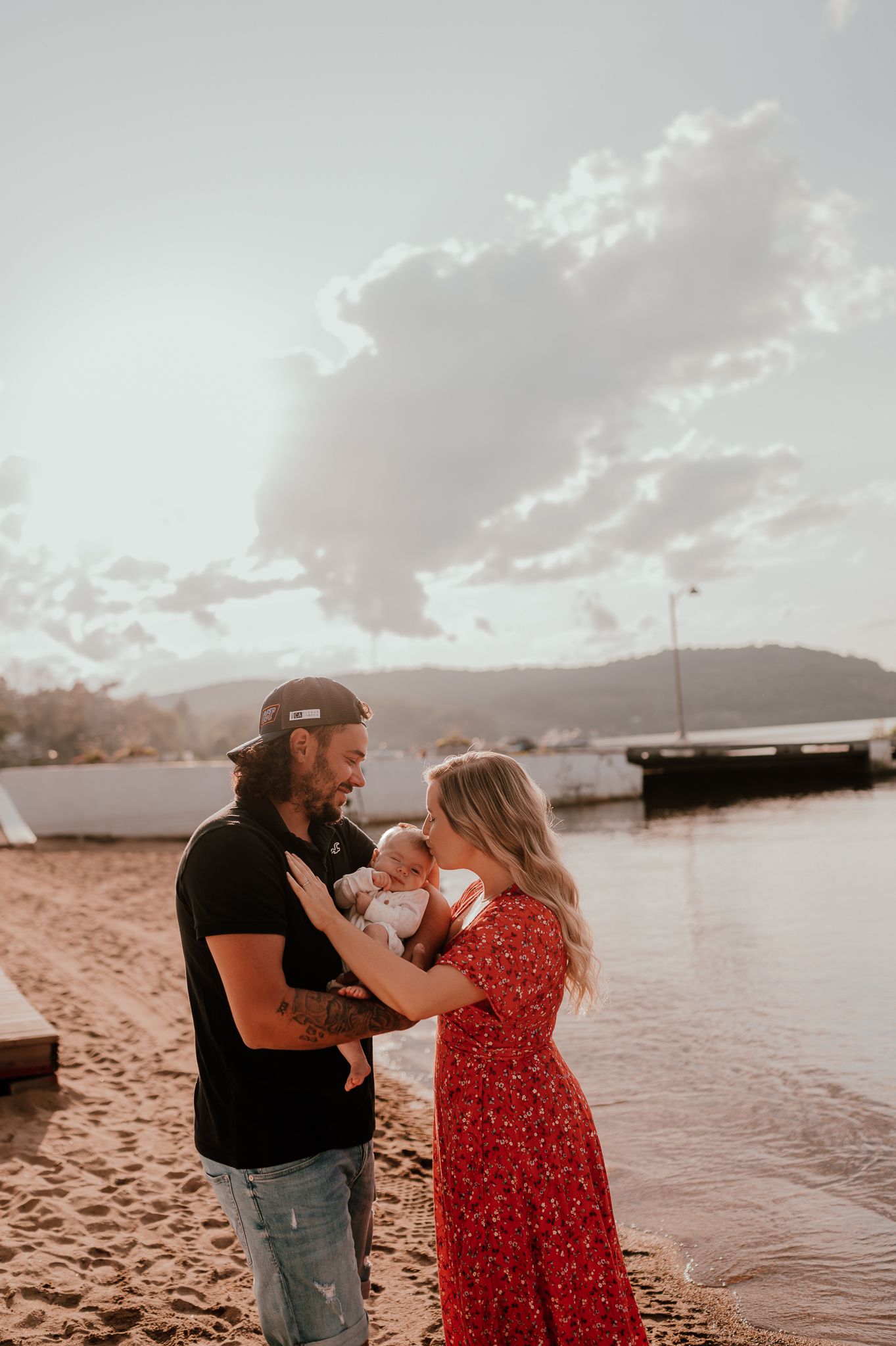 A couple holds their baby on a beach at sunset with boats in the background.