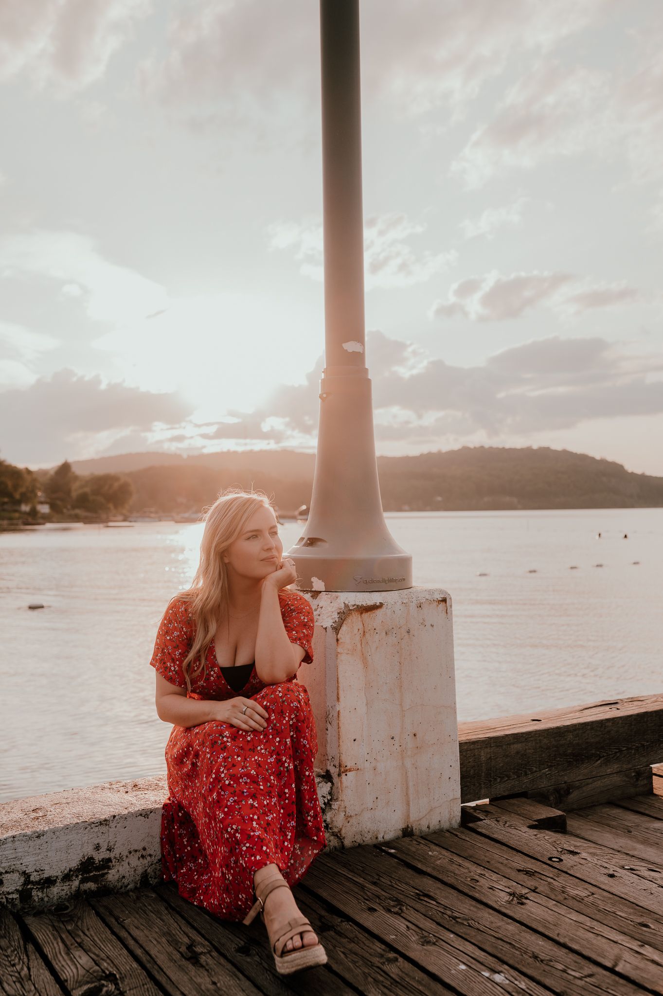 Woman in red floral dress sits thoughtfully on wooden pier during sunset.