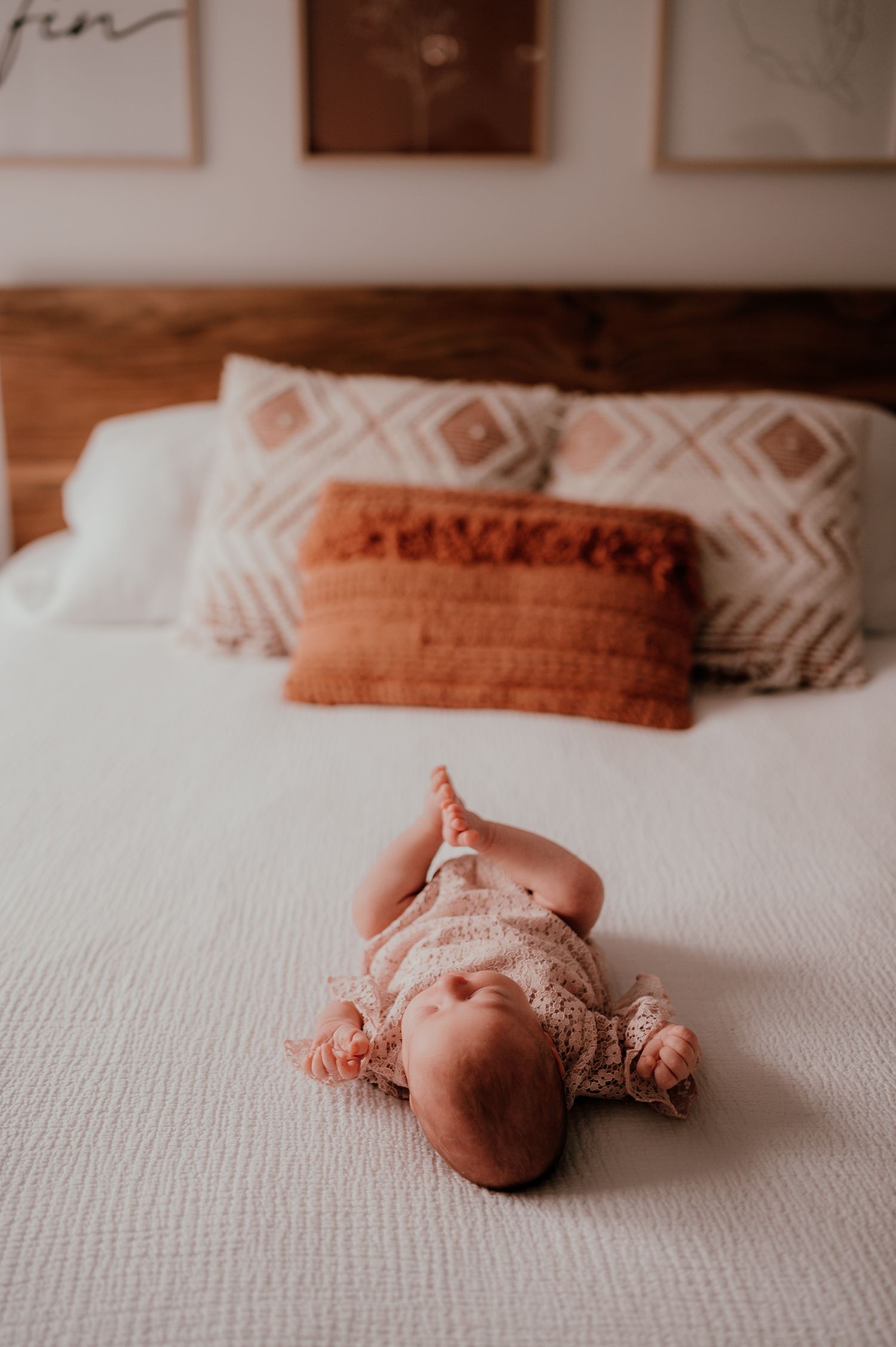 A baby wearing a lace outfit lies on a white bed with geometric patterned pillows.