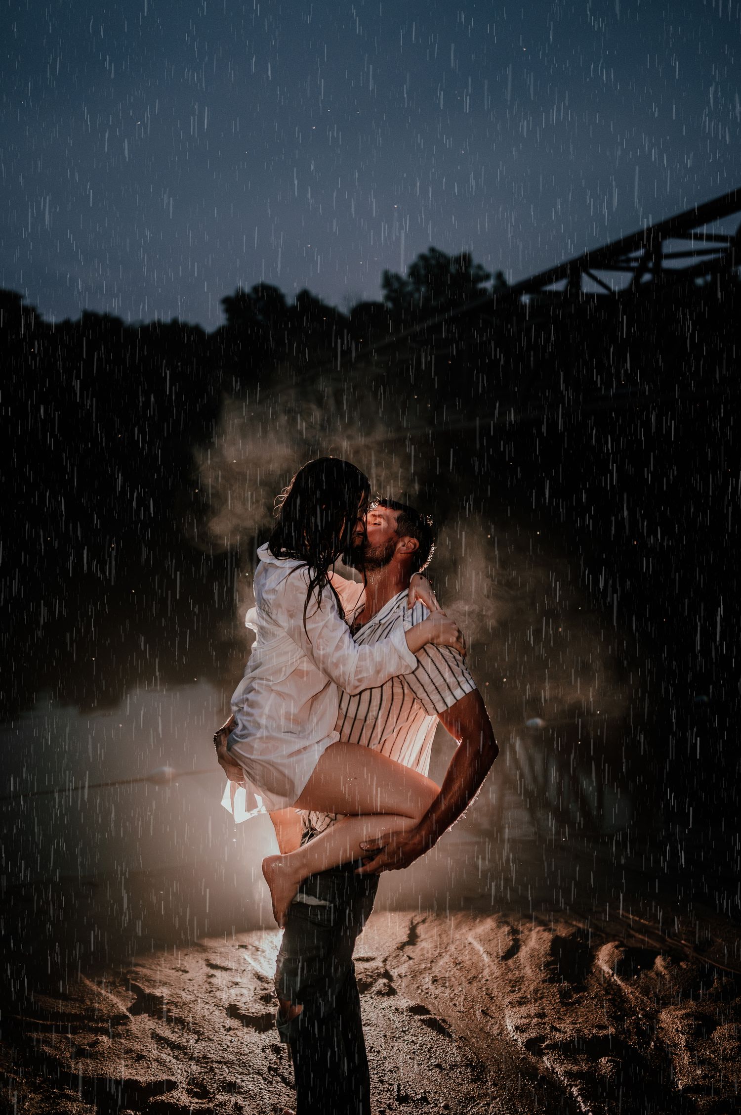 A romantic embrace in the rain with dramatic lighting and a bridge silhouette.