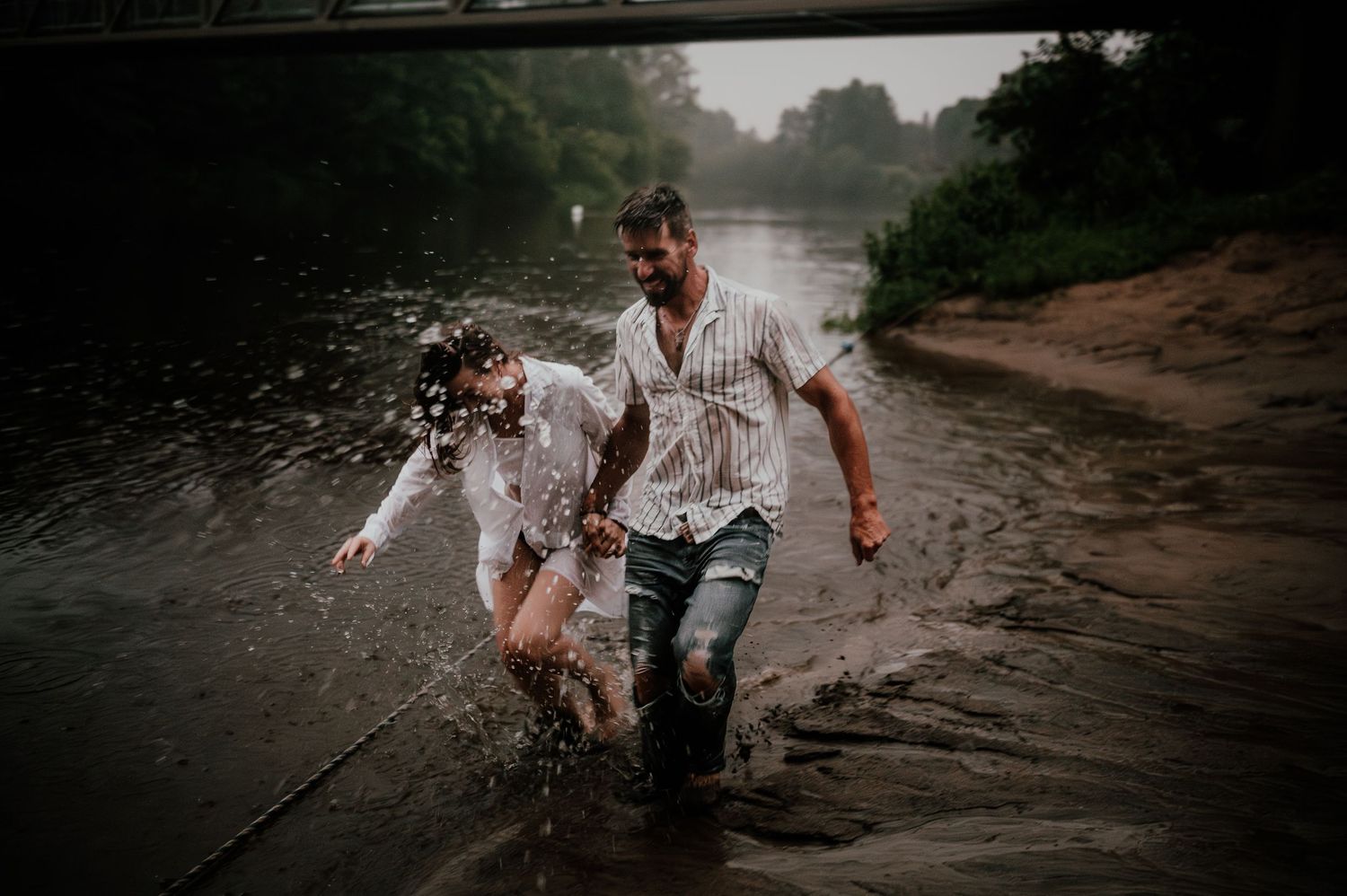 A couple splashing through water together during a moody outdoor photoshoot.