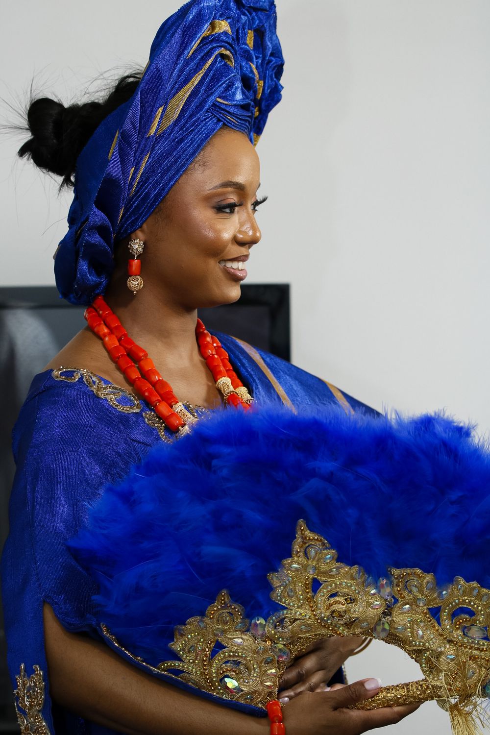 Traditional Nigerian bride wearing a royal blue gele headwrap and dress with gold embellishments and coral beaded necklace.