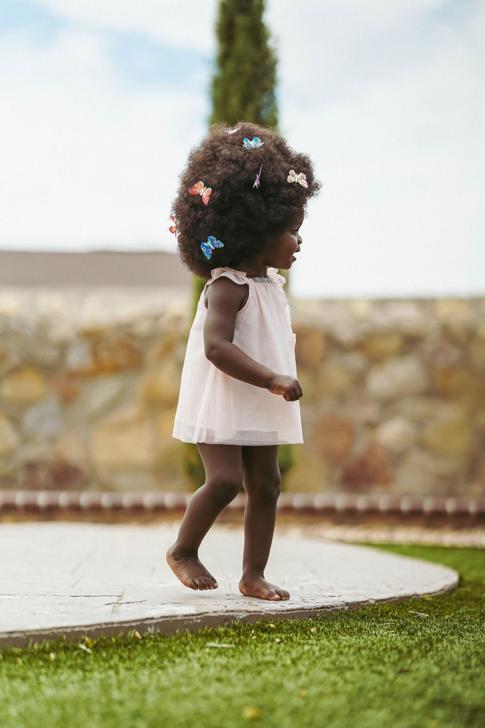 A small child in a white dress takes barefoot steps across a stone patio with palm trees in the background.