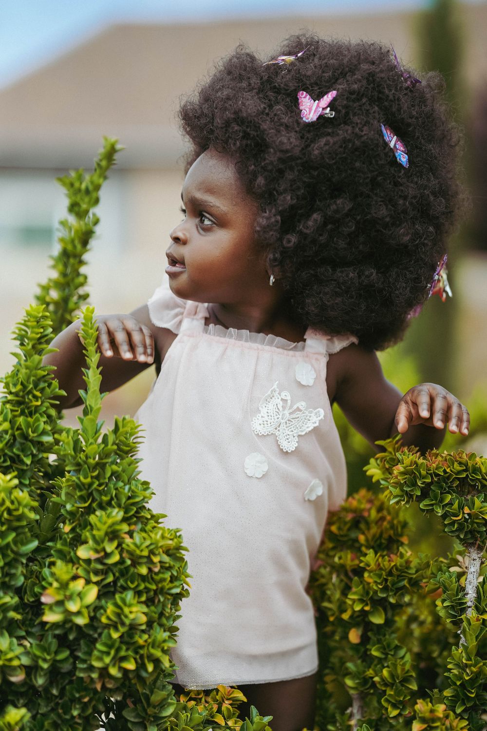 A child in white stands near green shrubs with decorative butterflies in their hair.