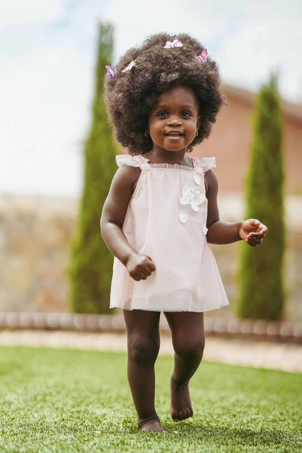 A joyful toddler dancing in a white summer dress on grass with small bows in their hair.