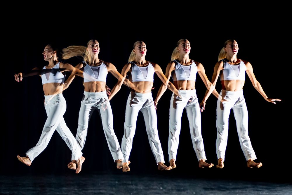 Dance group performs synchronized routine in matching white outfits against black background.