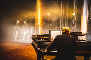 Piano player performing on stage in dramatic foggy lighting while silhouettes appear in background.