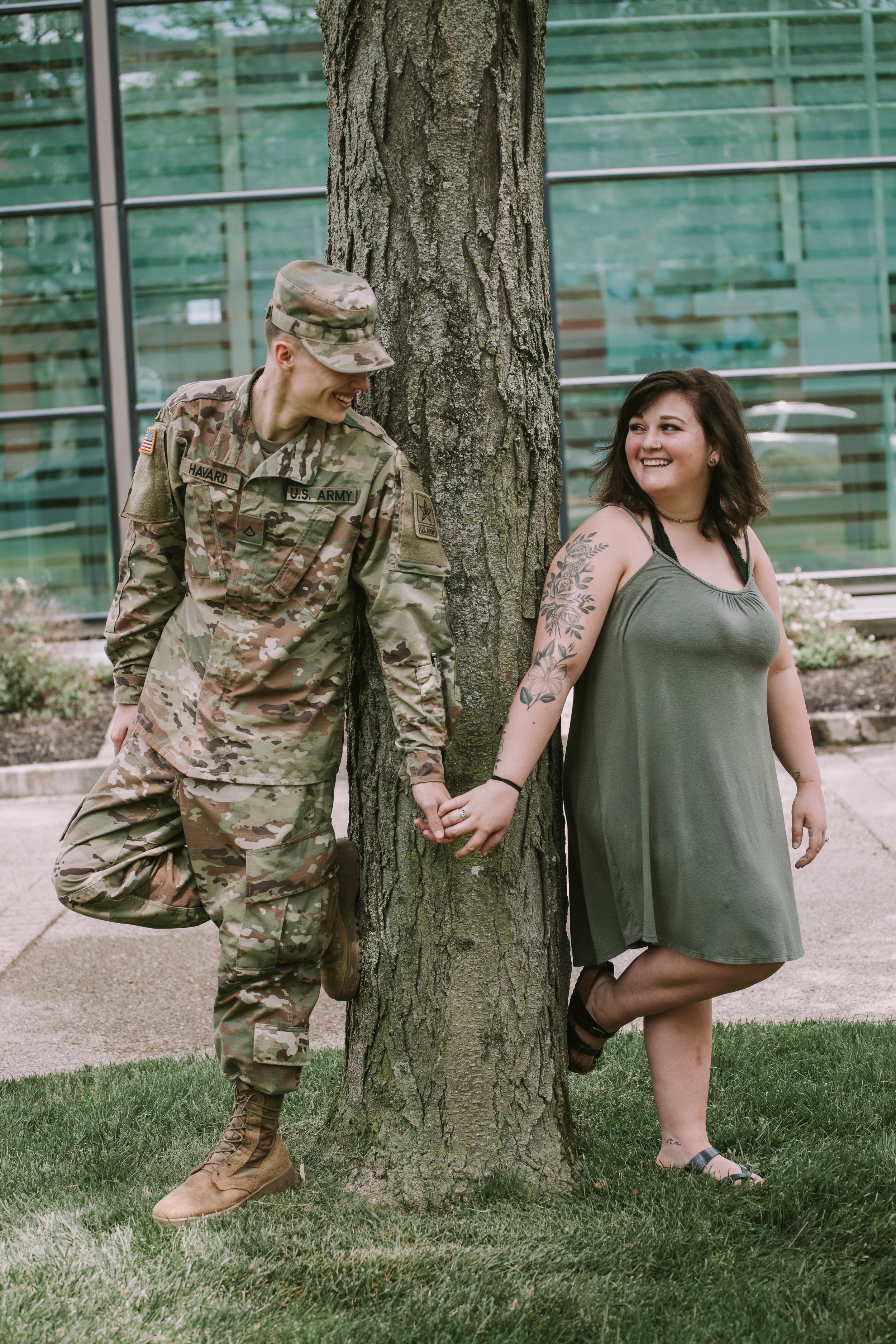 A couple poses playfully behind a tree trunk in an outdoor setting with stadium bleachers visible.