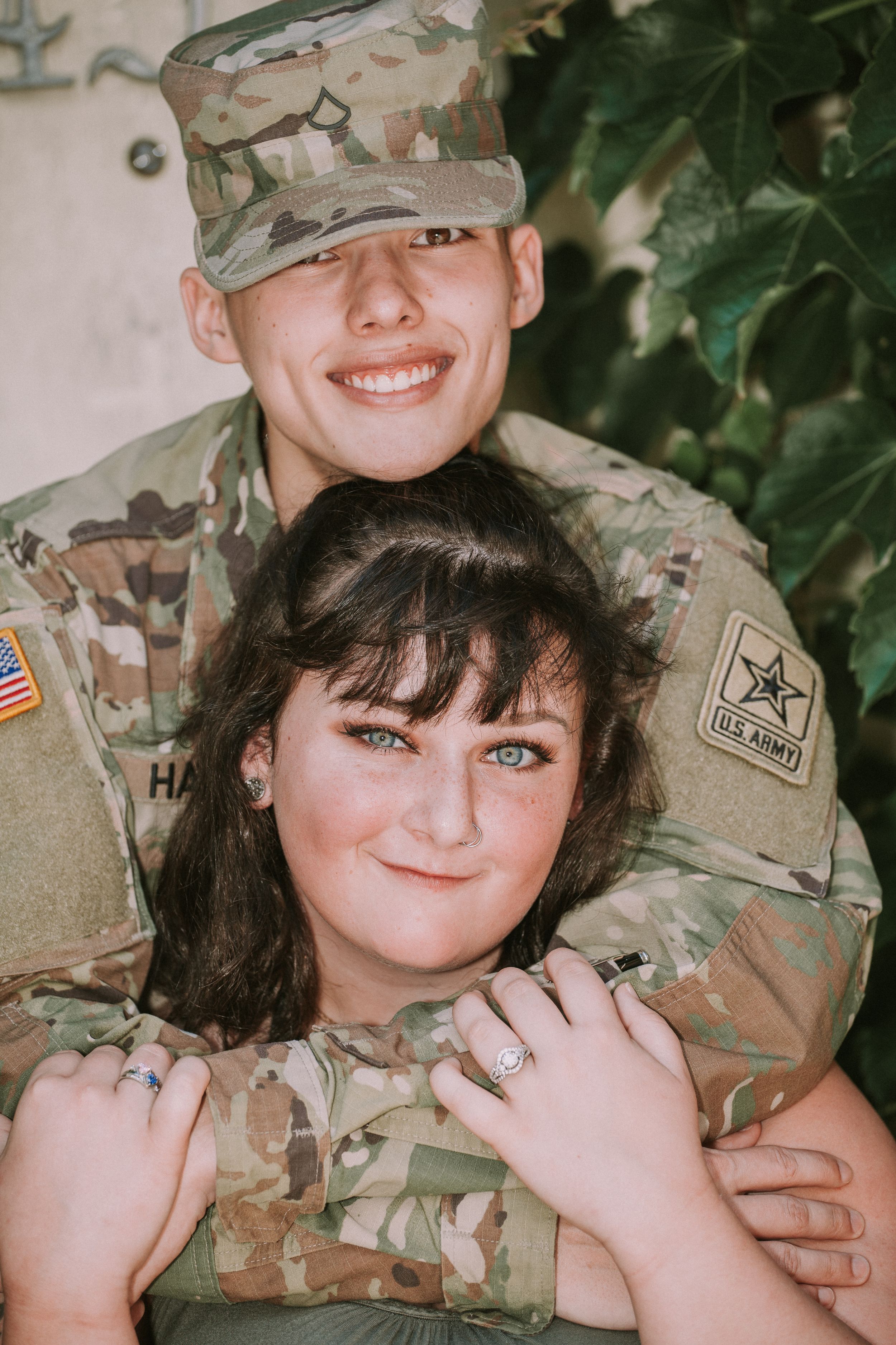 A couple poses together in military camouflage uniform and casual attire showing affection and smiles.
