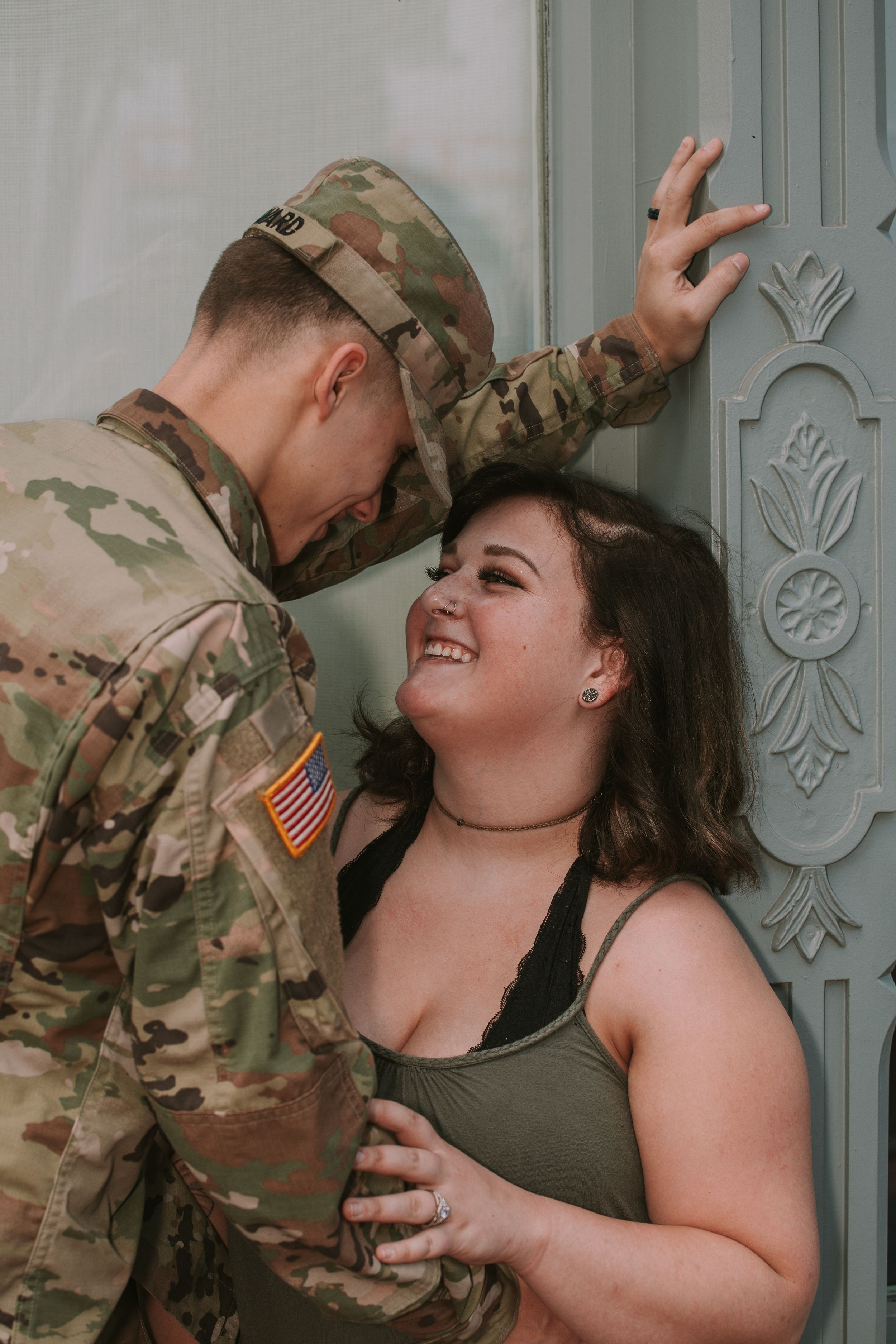 A couple shares an intimate moment against a decorative door, one in military fatigues.