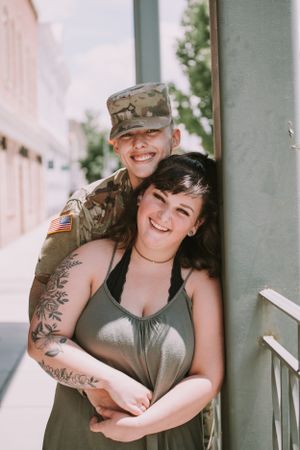A couple embraces and laughs together on a porch, one wearing military attire.
