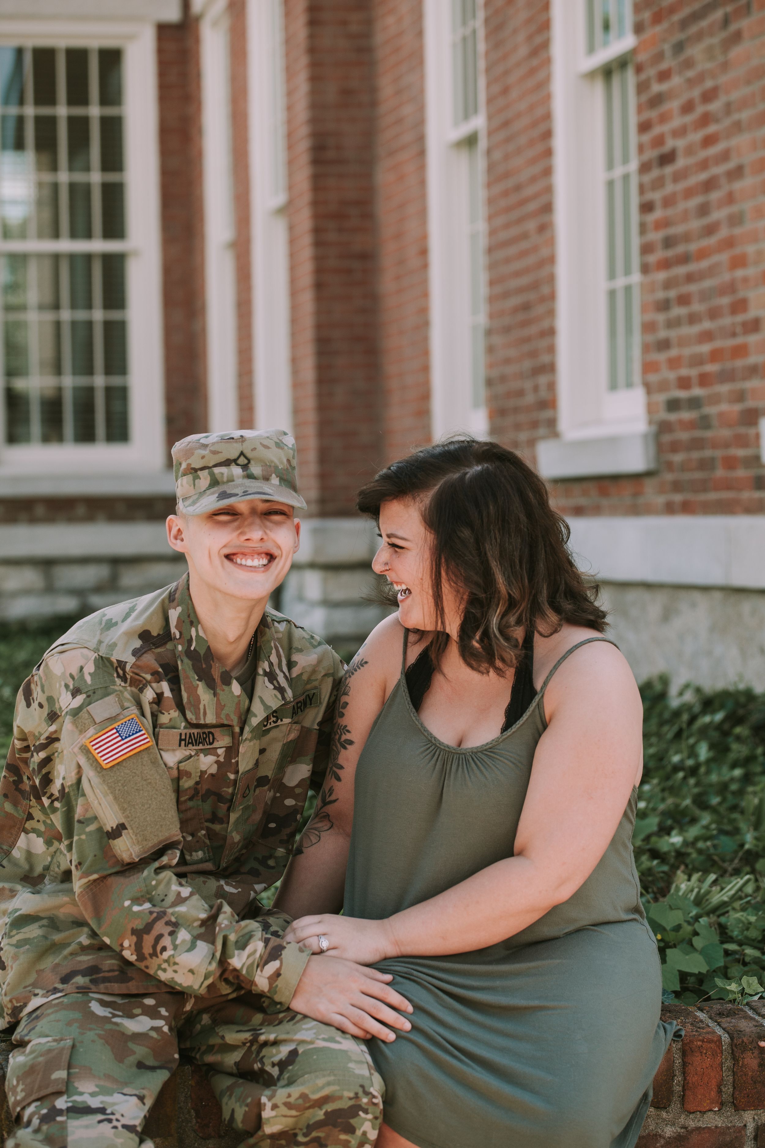 A couple shares a playful moment together against a brick building backdrop, one wearing military camouflage.
