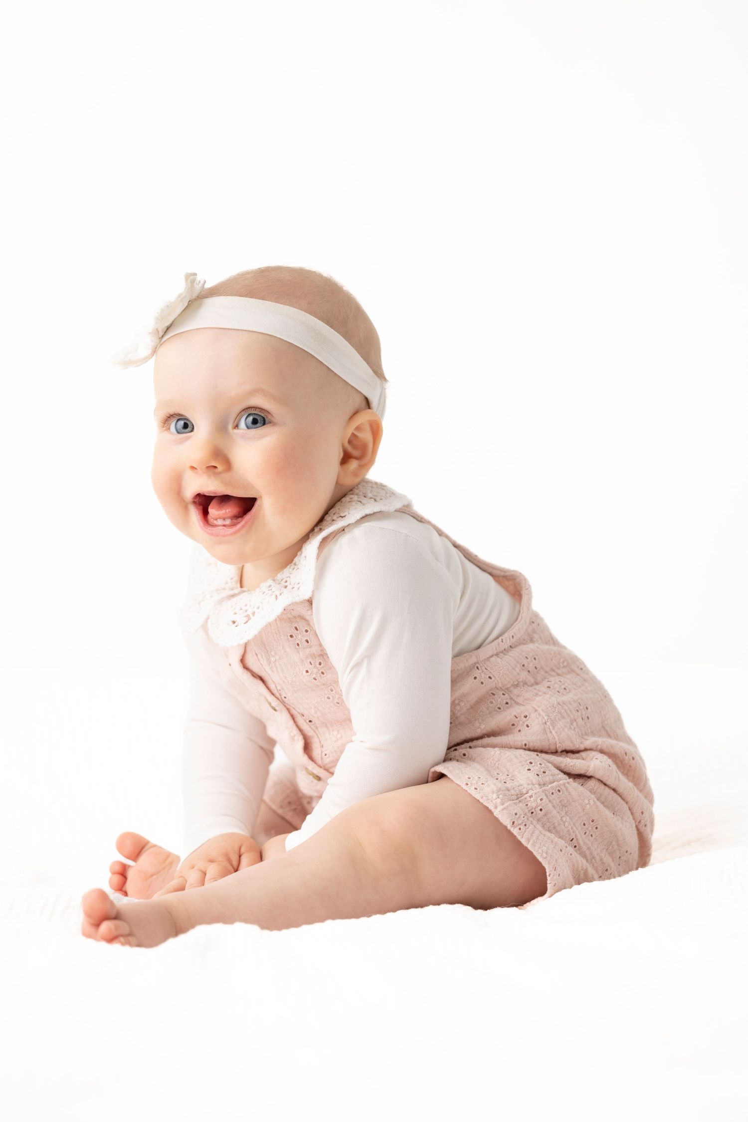 Smiling baby wearing white headband and pink romper sitting on white background.