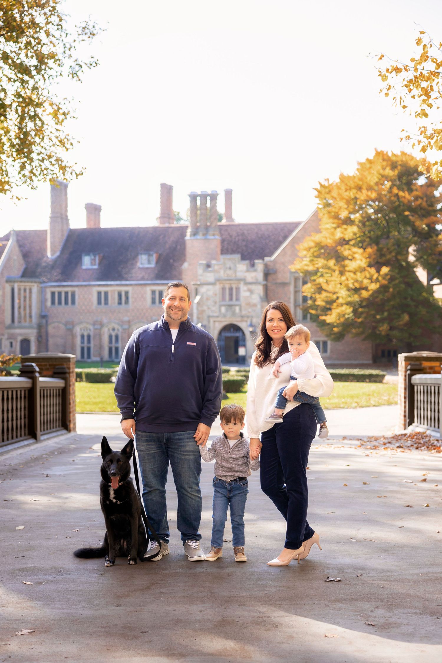 Family and their black dog pose together on pathway with grand mansion in background during autumn.