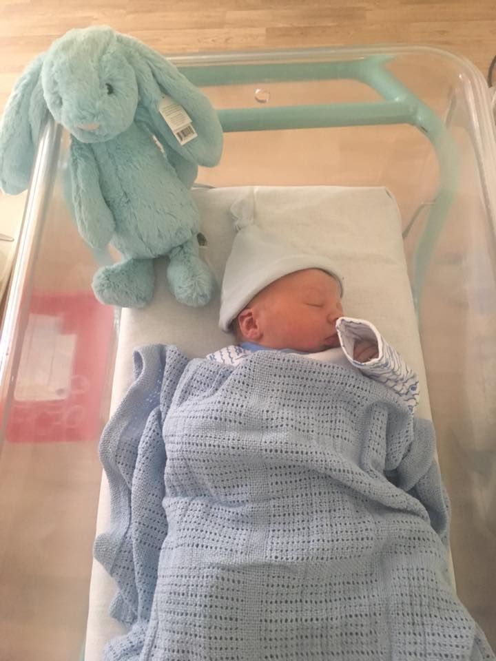 Sleeping newborn in blue blanket with mint green stuffed bunny in hospital bassinet.