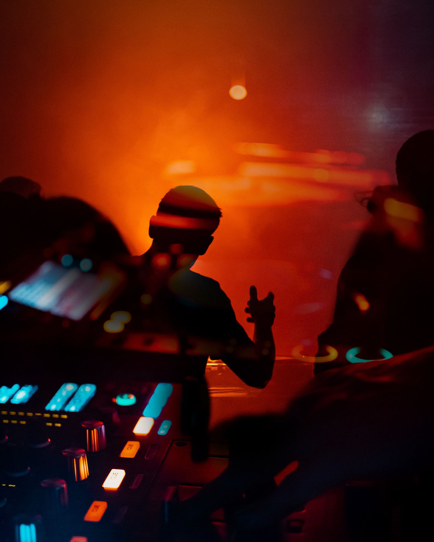 DJ silhouettes performing against vibrant orange sunset backdrop with equipment illuminated in blue.