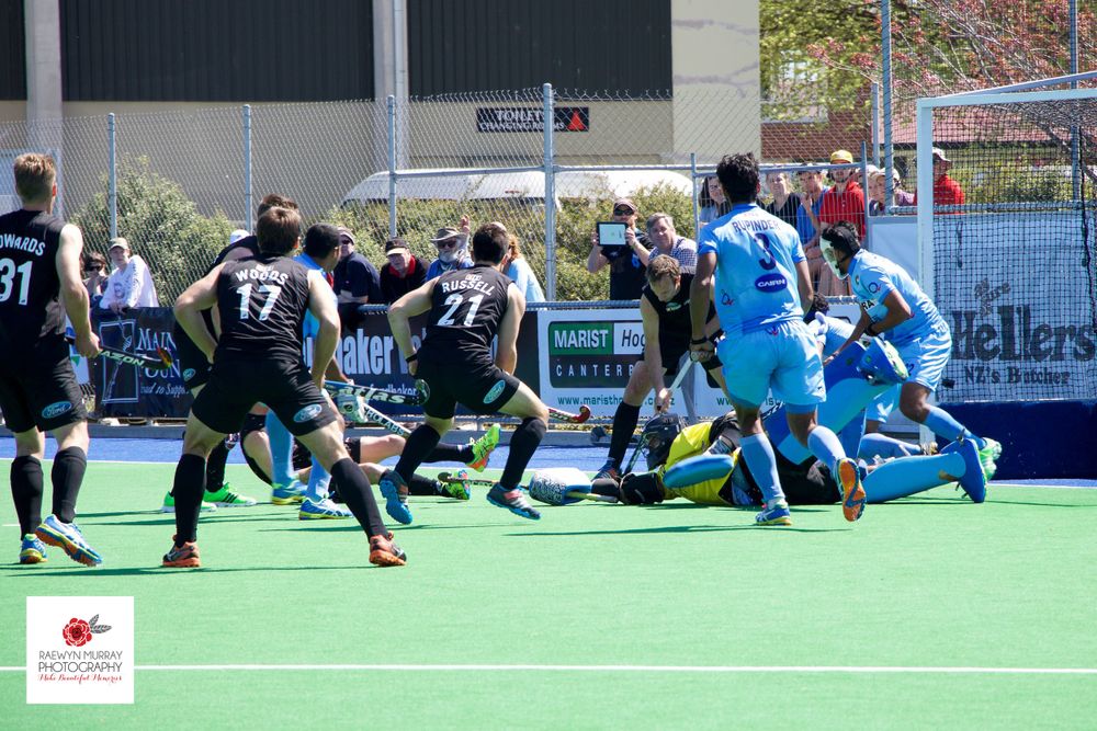 Field hockey players in black uniforms compete against players in blue during an intense match on artificial turf.