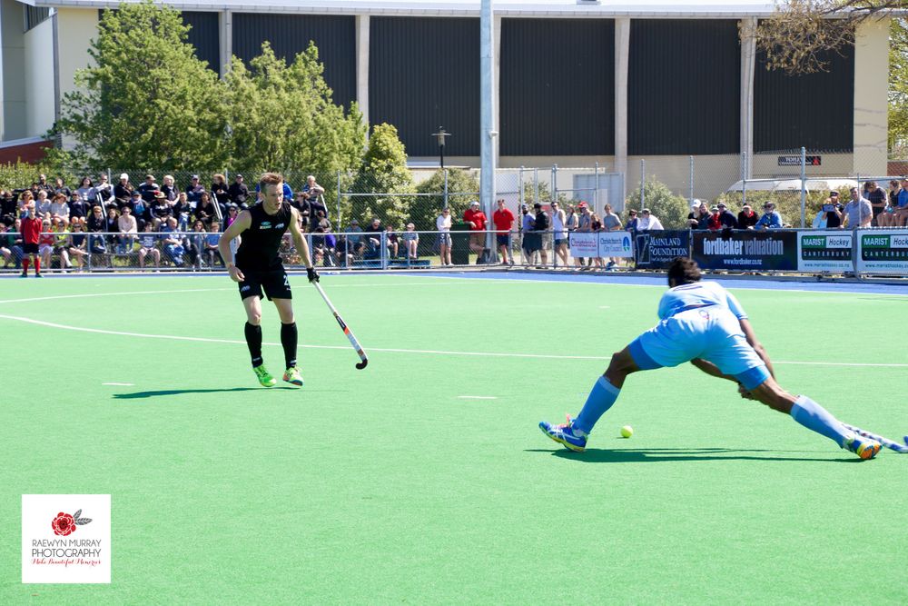 Two field hockey players face off on artificial turf with spectators watching from the sidelines.