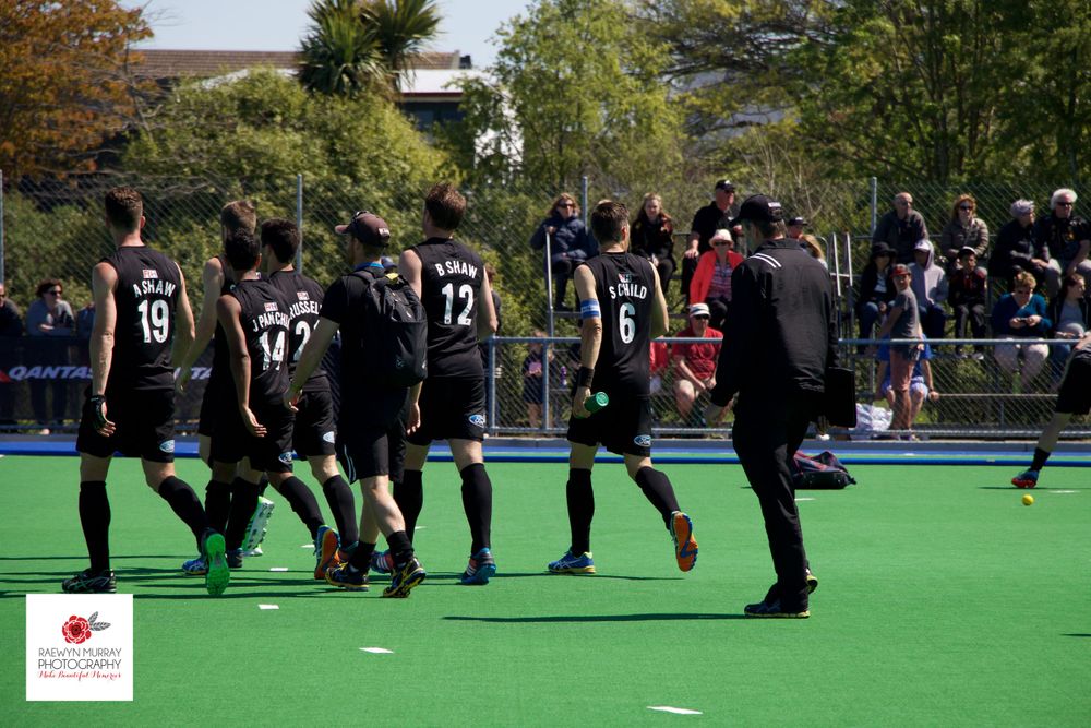 Team in black uniforms huddles together on field hockey pitch during game break.