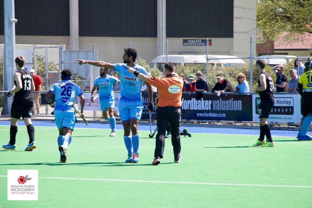 Field hockey match officials signal during gameplay on artificial turf.
