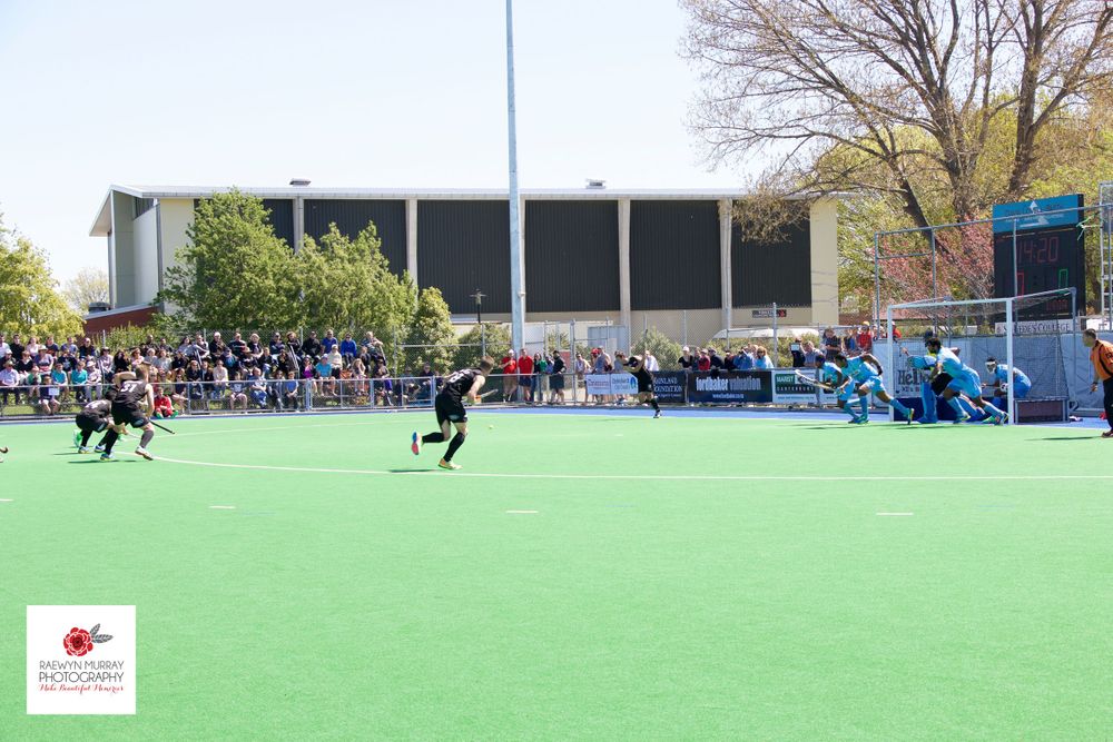Field hockey match in progress on artificial turf with spectators watching from sidelines.