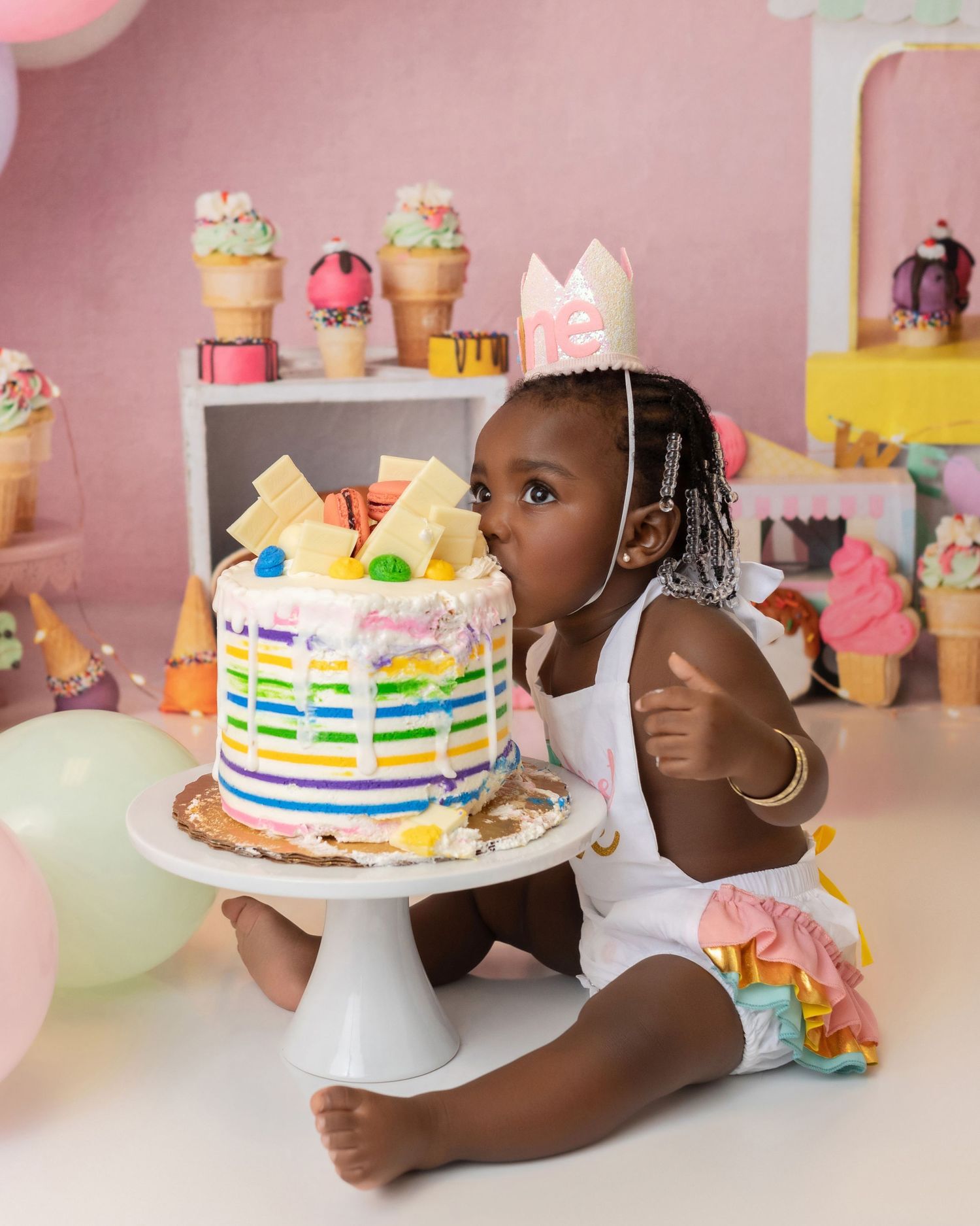 A festive rainbow-striped birthday cake sits on a white pedestal stand against a pink backdrop with party decorations.