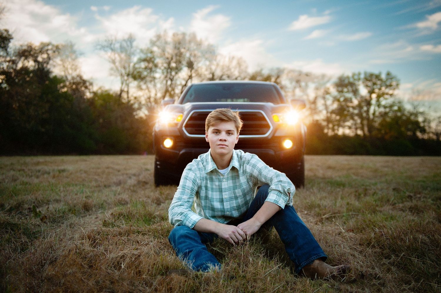 Young person in plaid shirt sitting on ground with vintage truck headlights glowing at sunset.