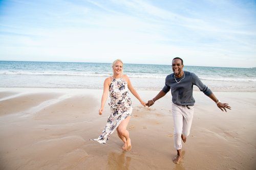 A couple runs joyfully hand in hand along a sunny beach shoreline.
