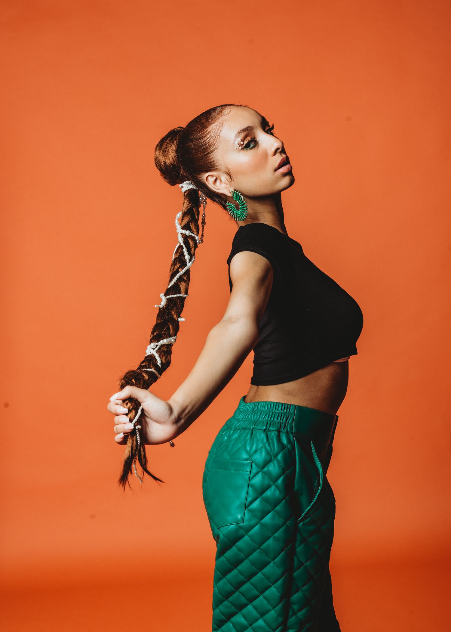 Fashion model in black crop top and green quilted pants poses against orange background with braided hairstyle.