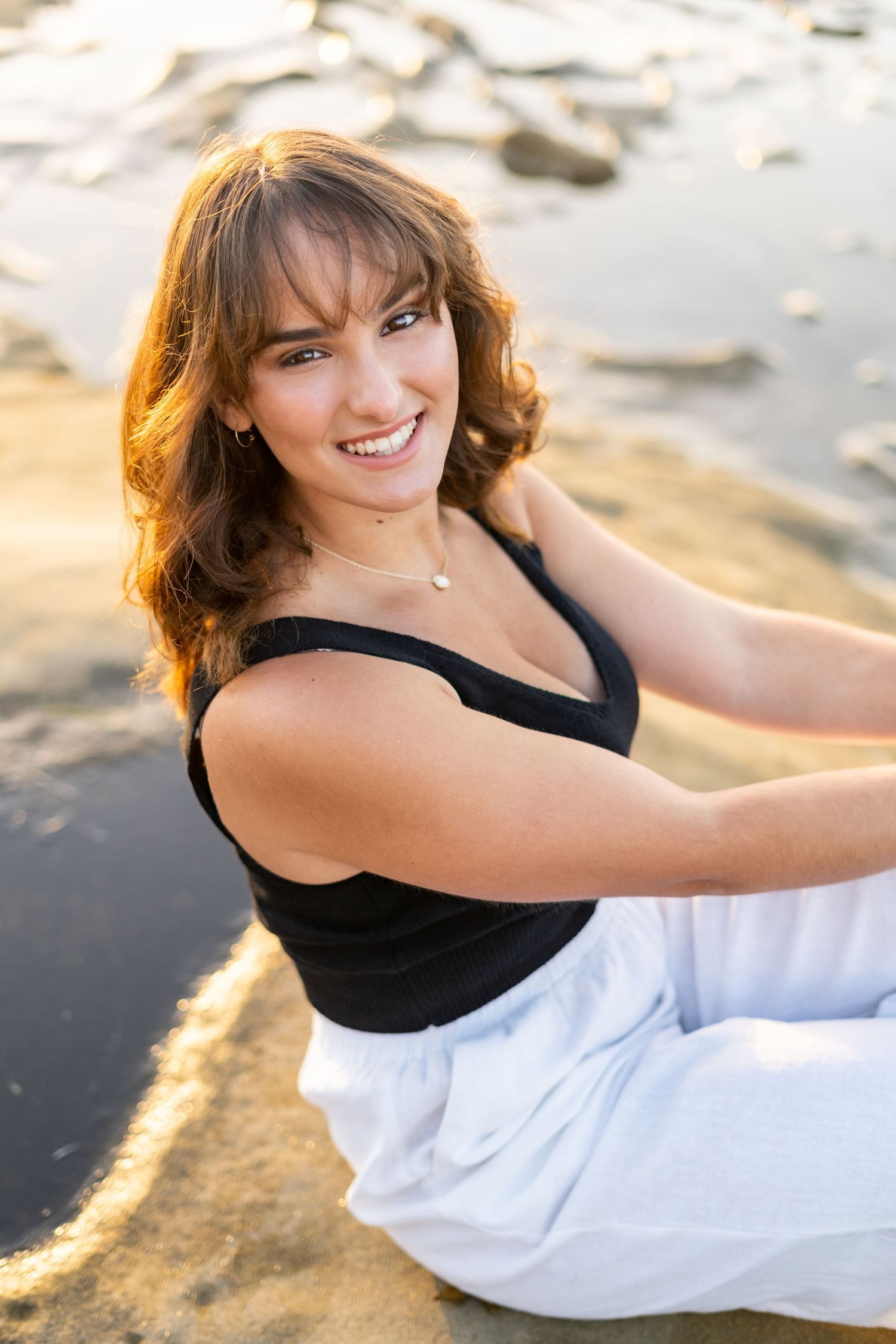 A high school senior in black tank top and white pants sitting on beach at sunset.