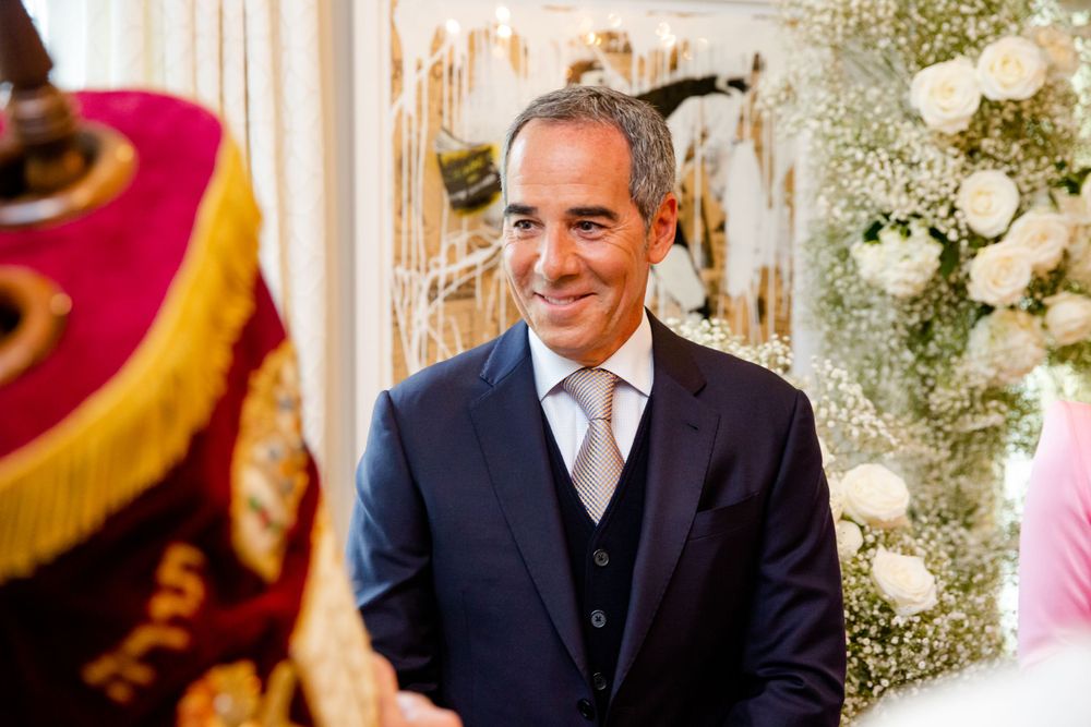 Guest in navy suit smiles during ceremonial presentation with white floral arrangements.