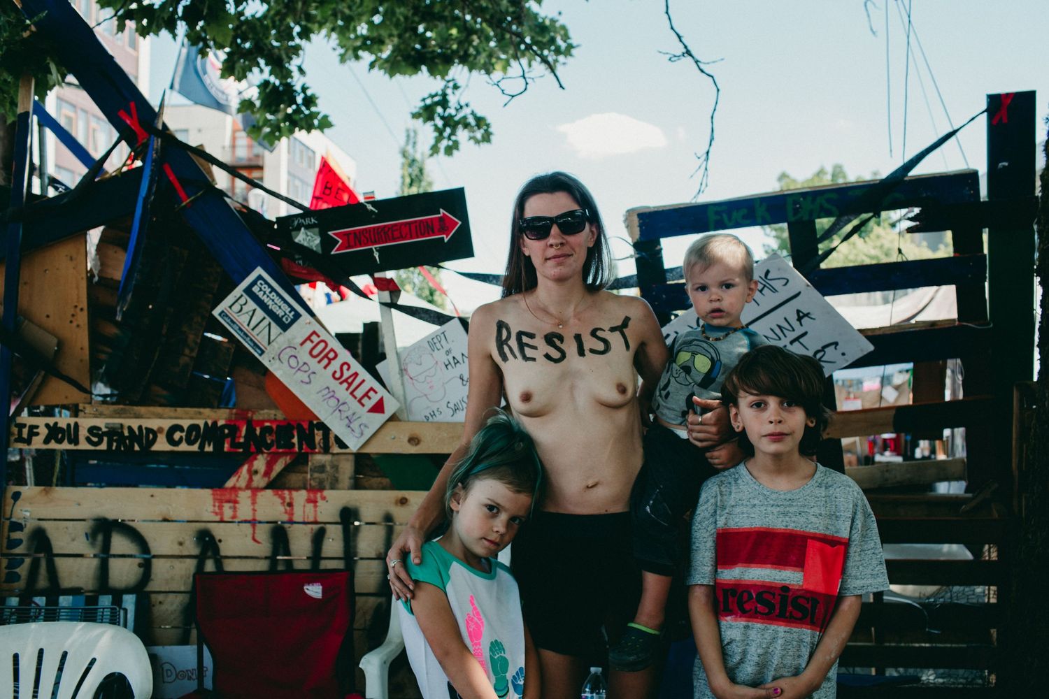 shirtless parent with RESIST painted on chest stands with three children at Occupy ICE portland oregon