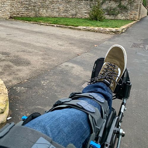 A person's leg in jeans and sneakers propped up in a wheelchair outdoors.
