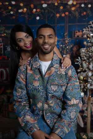 Couple poses in front of Christmas tree with decorative holiday backdrop.