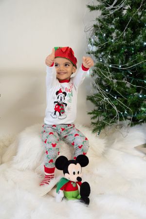 Child in Mickey Mouse Christmas pajamas playing by decorated tree with plush toy.